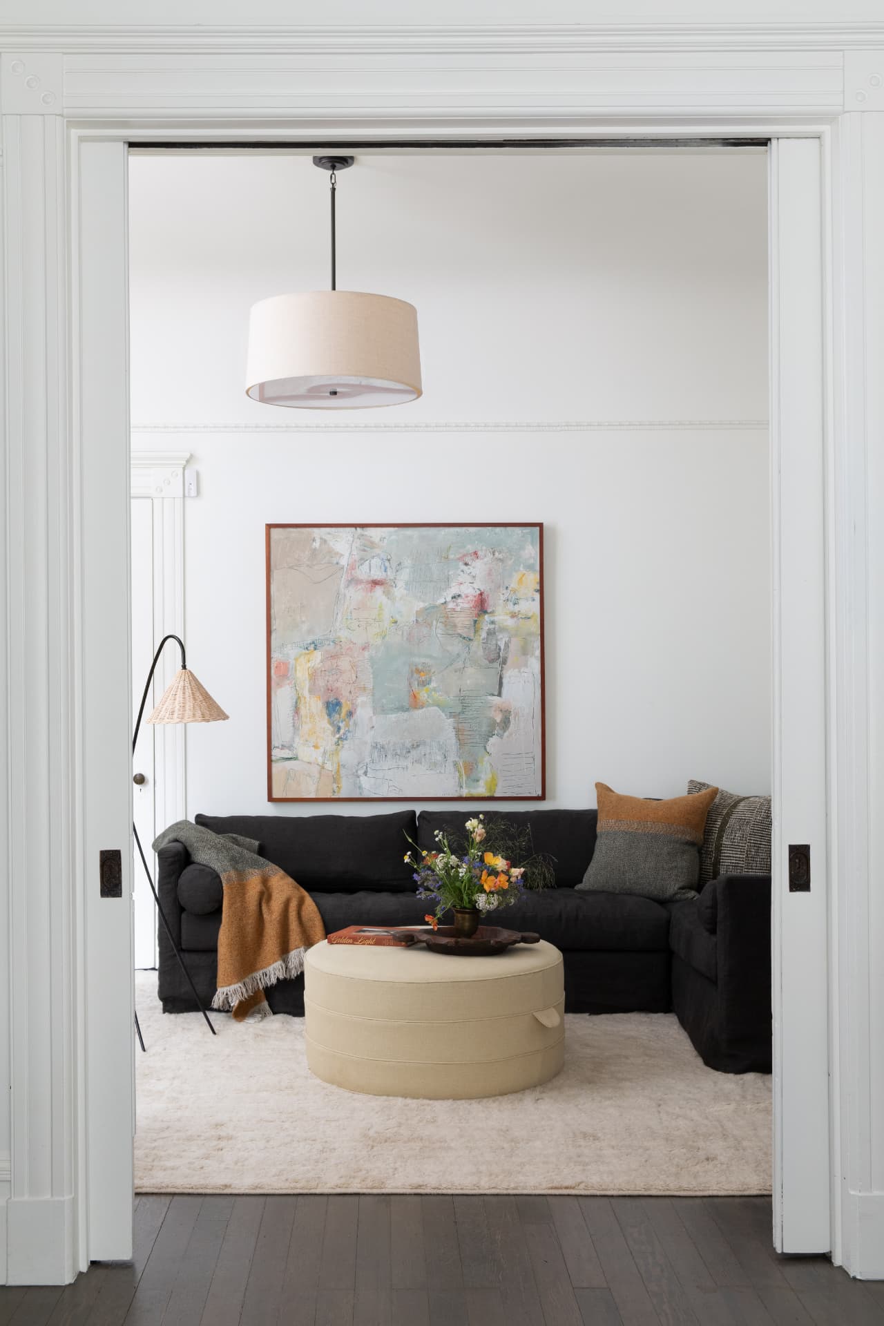A view through pocket doors into a living room with a dark sofa, abstract artwork, and a linen drum pendant