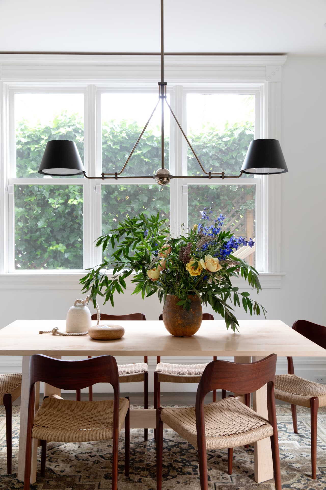 A light-filled dining room with a wooden table, cane-back chairs, and fresh flowers beneath a black chandelier